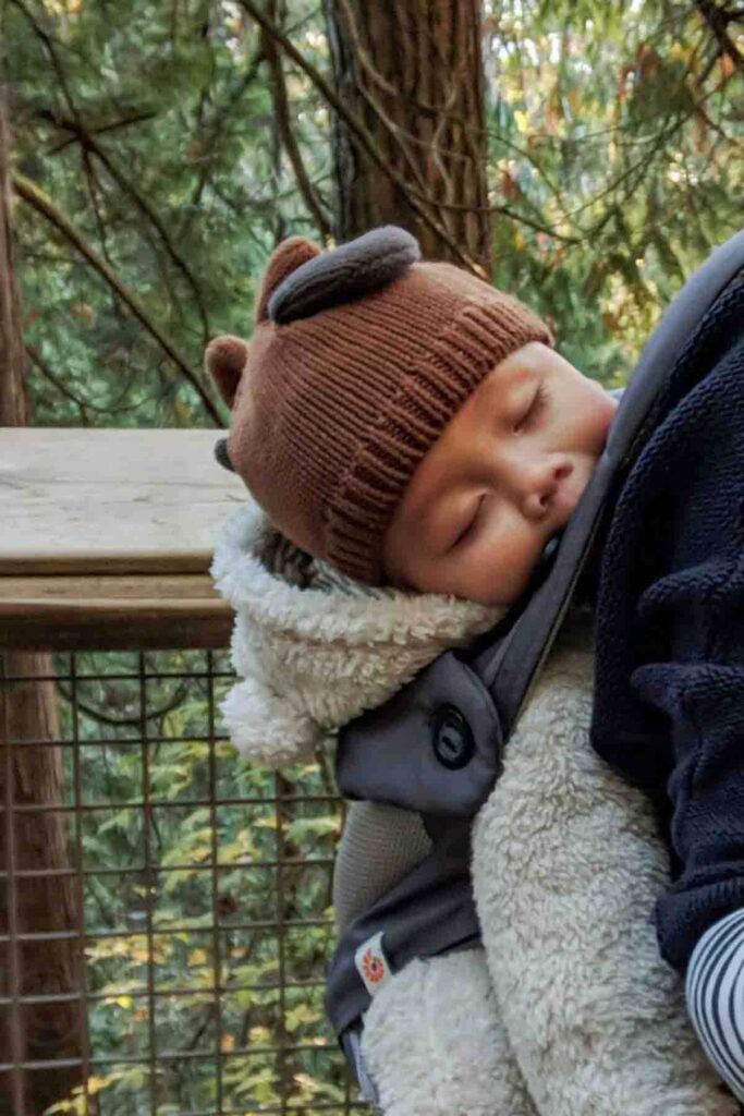 Baby wearing a fuzzy bear suit asleep on their parent’s back in an Ergo carrier during a Vancouver trip wearing a knit hat demonstrating baby nap on the go and practical baby sleep travel tips for sightseeing days.