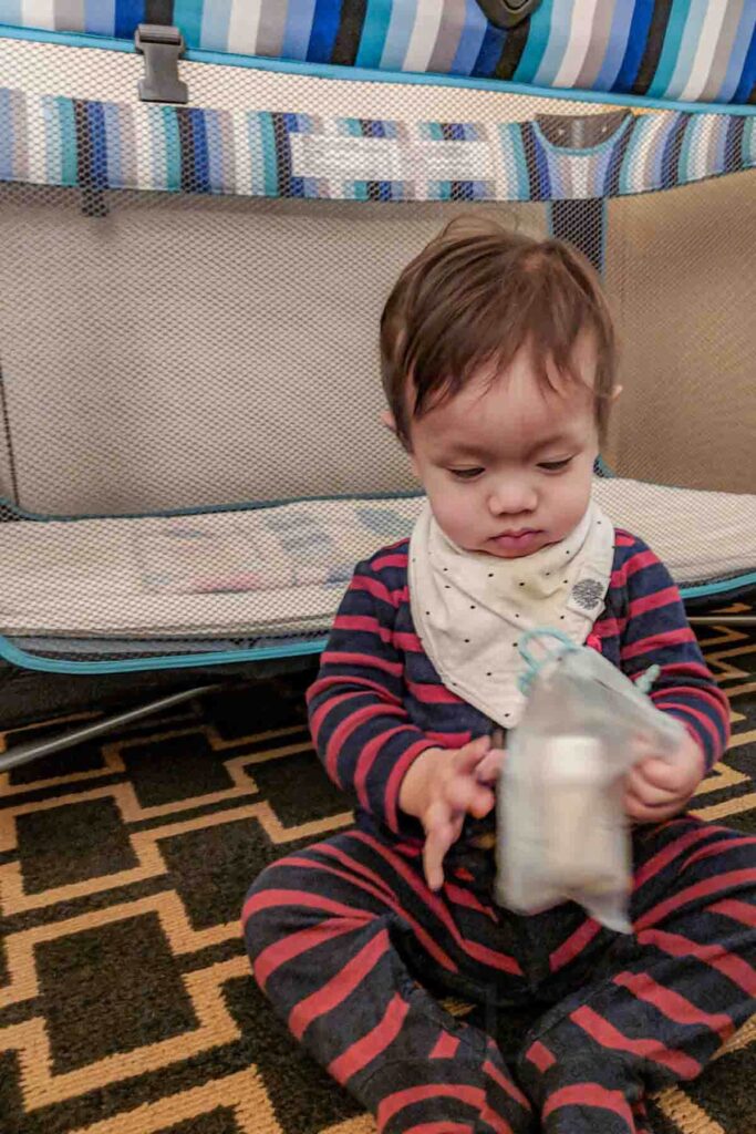 Baby wearing red and navy striped pajamas wearing a white modern bib sitting on the floor in front of a mesh sided hotel crib while holding a small pouch.