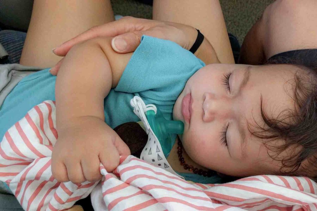 Baby sleeping on a train gripping a striped nursing cover with a green soothie pacifier next to him, illustrating baby sleep while traveling.