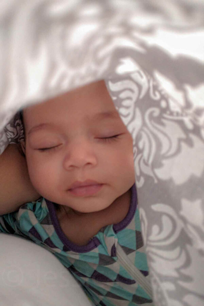 Newborn sleeping peacefully underneath a patterned damask gray and white nursing cover creating a darkened space that works as a baby blackout solution travel option and portable sleep solutions for baby.