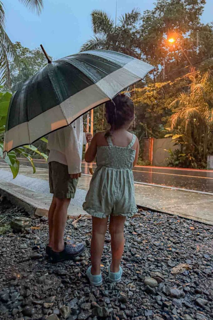 Two young children stand under a large umbrella in the rain at dusk while streetlights glow and palm trees sway in the background in Bocas del Toro, Panama. The moody evening setting visually represent jet lag and how travel can disrupt sleep routines.