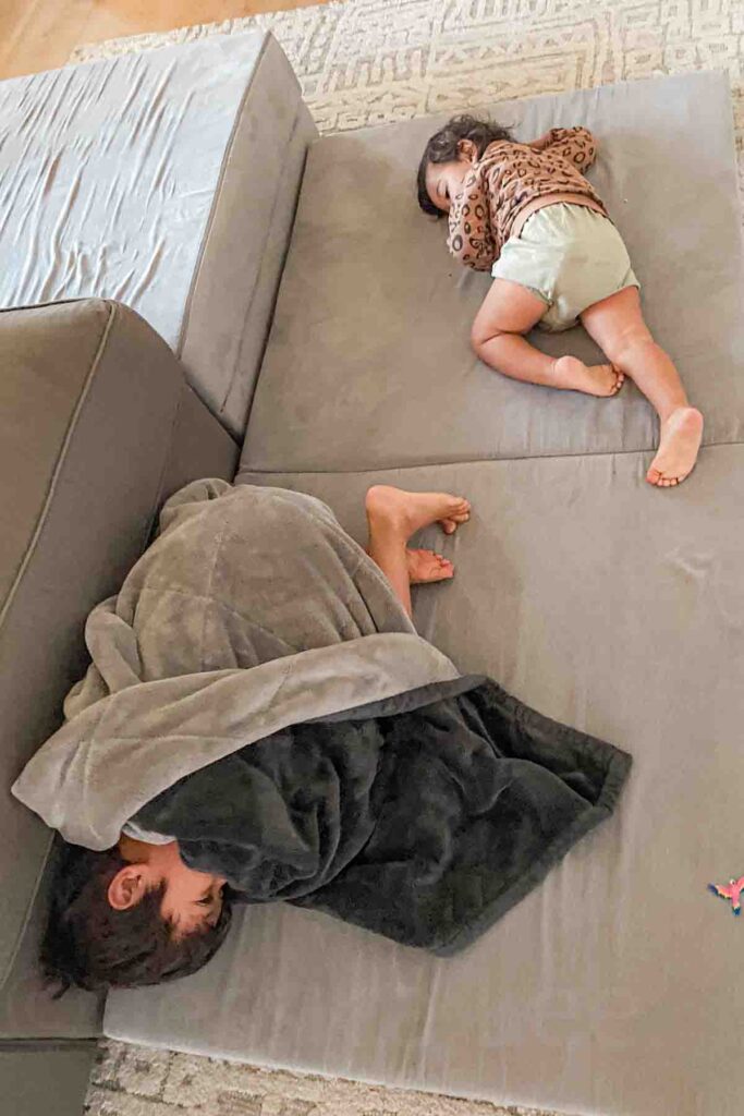 Two children nap on a tan Nugget couch in the middle of the day, one curled under a dark blanket and the other toddler sleeping without one. The daytime sleeping scene reflects jet lag in children and how adjusting back can be tough.