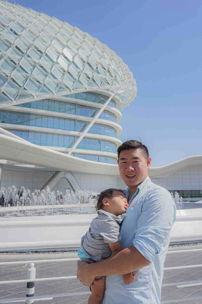 A father holds a sleeping young toddler outdoors in bright daylight in front of a modern glass building and fountain at the F1 Grand Prixe track in Abu Dhabi. The child resting in transit highlights jet lag with a baby and how to adjust to a new time zone while traveling.