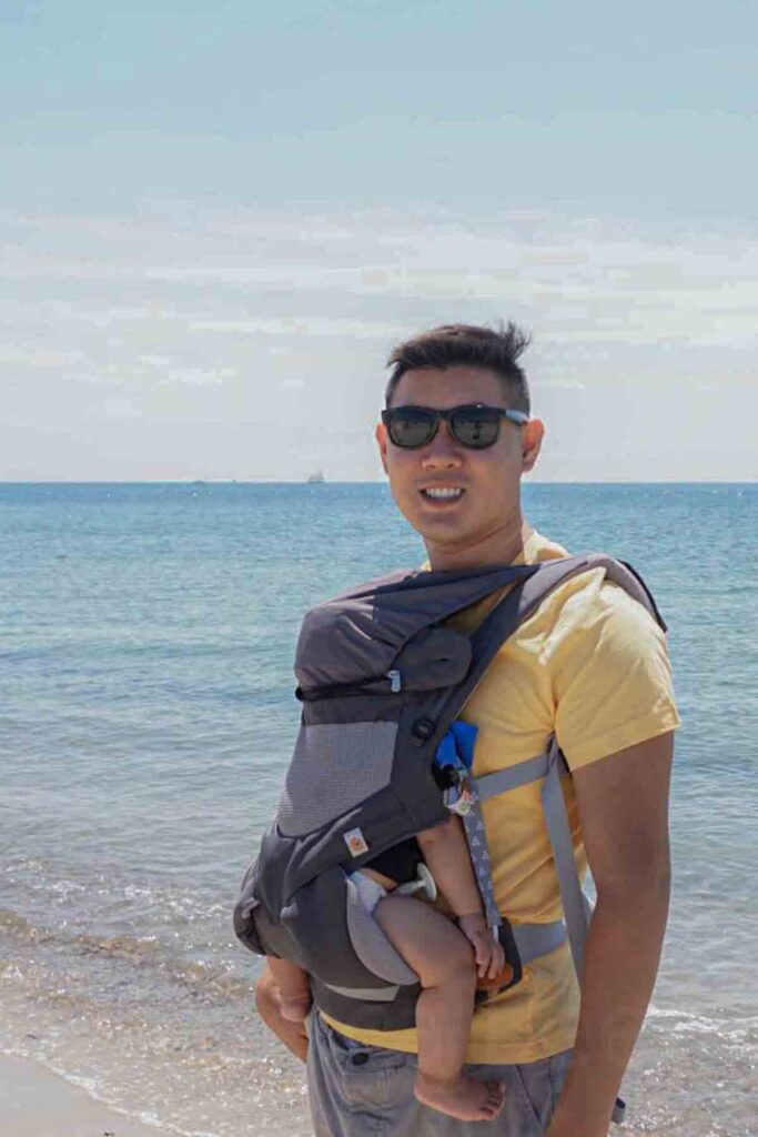 Dad standing by the ocean wearing a baby in a front carrier while the baby sleeps against his chest. Getting a baby to sleep while traveling is easier with hands free carriers that support baby sleep on the go during beach walks and outdoor adventures.