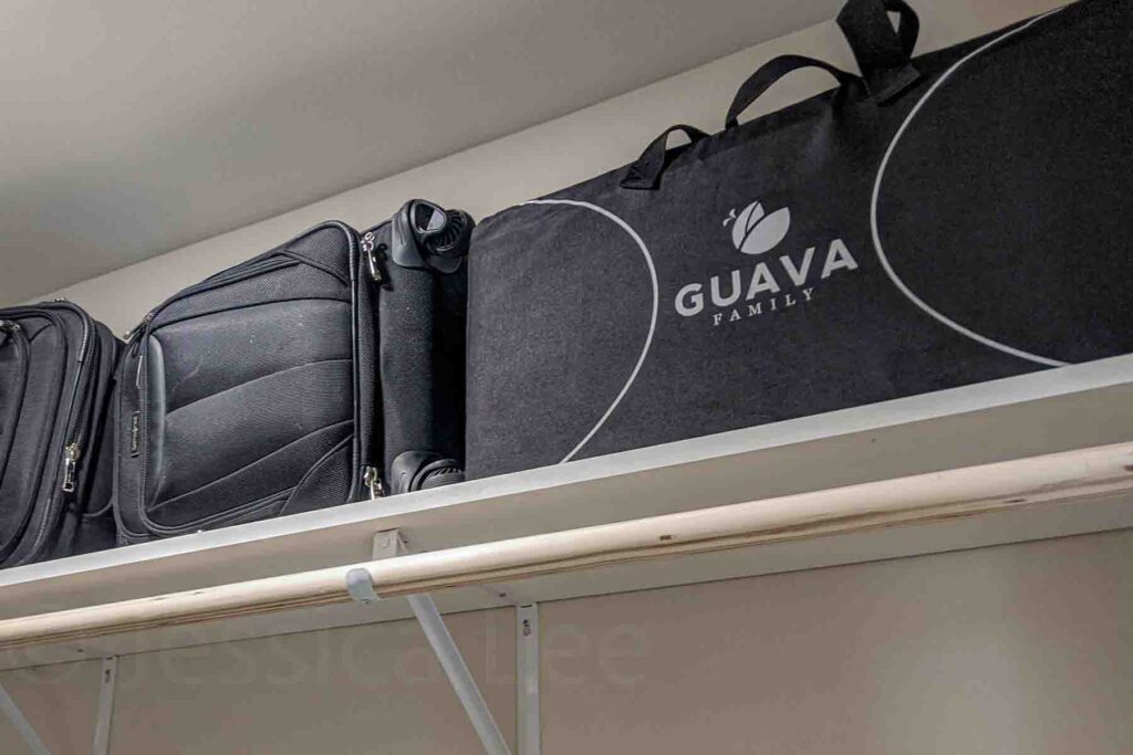 Guava Lotus travel crib packed in its black bag stored on a closet shelf along with two black small suitcases showing how compact and portable this baby sleep solution is for traveling with a baby.