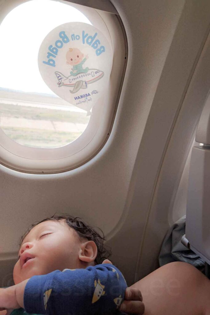 Lap infant asleep during a plane ride next to an airplane window with a Baby on Board sticker illustrating the perfect example of a baby sleeping while traveling in real life.