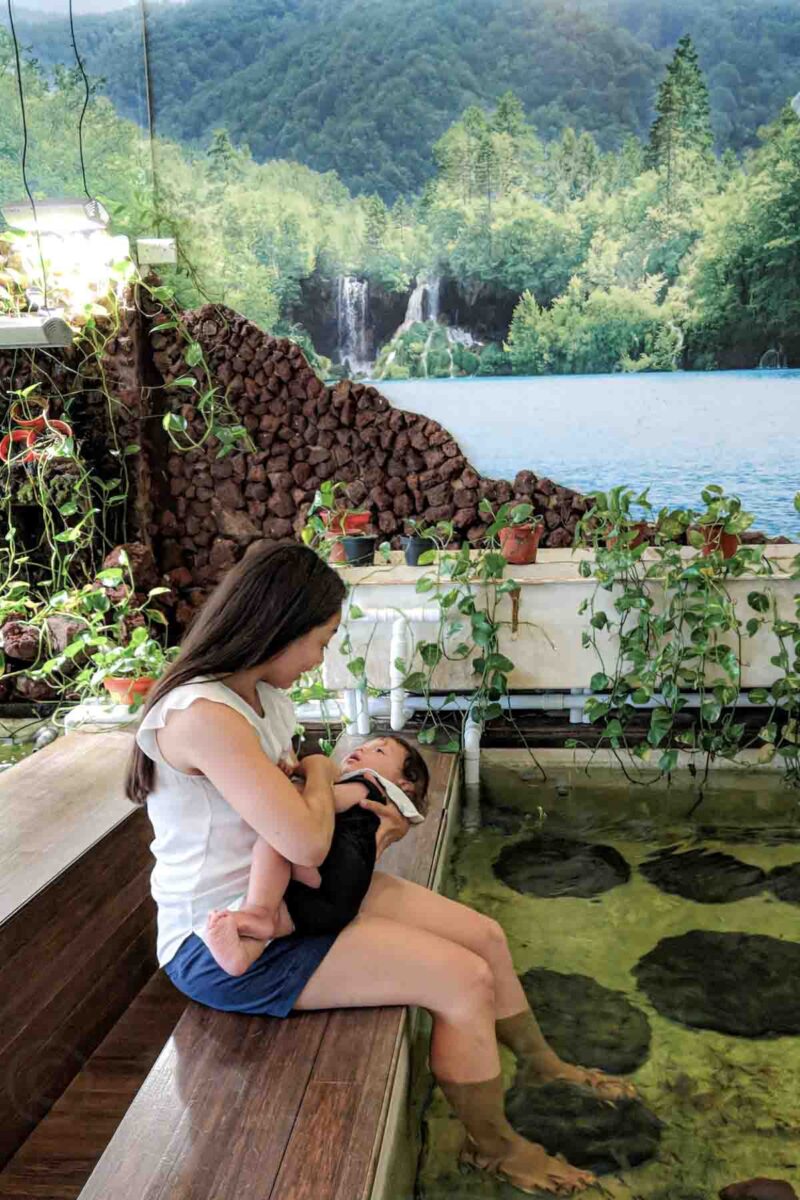 Mother sitting at the edge of a fish spa pool in Singapore holding her baby preparing to nurse him while small fish swim around their feet, showing a relaxed break while still feeding baby during a trip.
