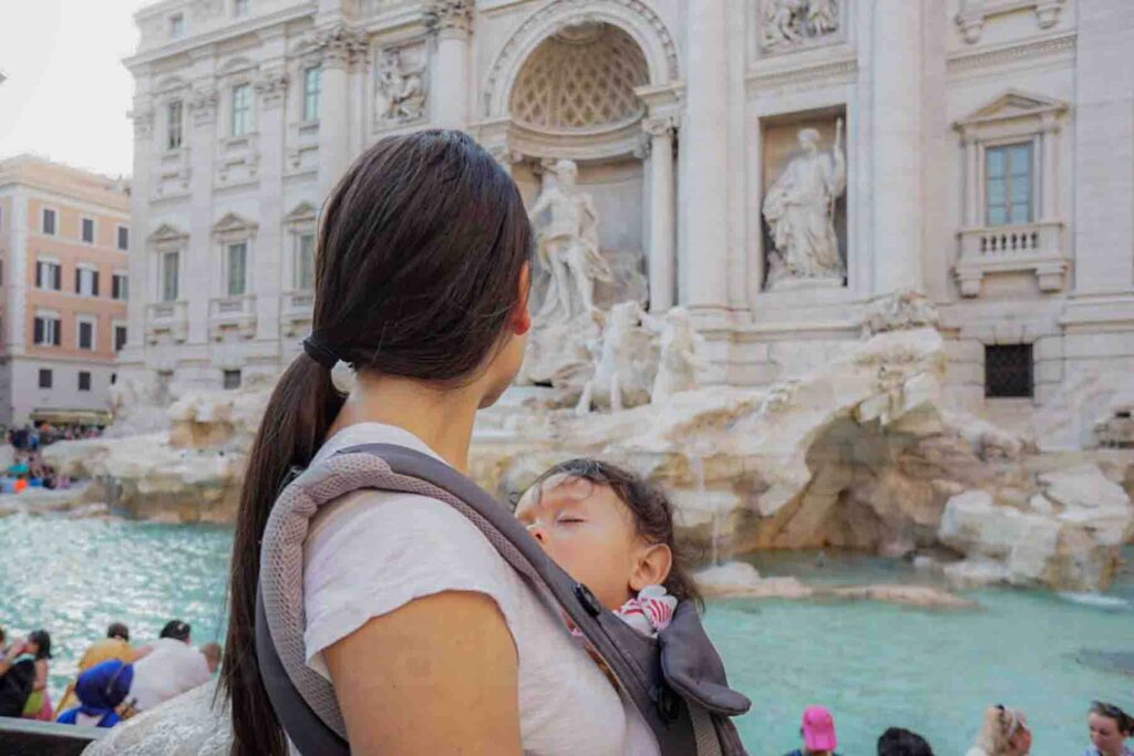 Mom standing in front of the Trevi Fountain in Rome, Italy while her baby sleeps tucked into a soft structured Ergobaby carrier against her chest. This is a real life example of having a baby sleep while traveling in busy tourist spots using baby travel sleep gear to keep naps consistent.