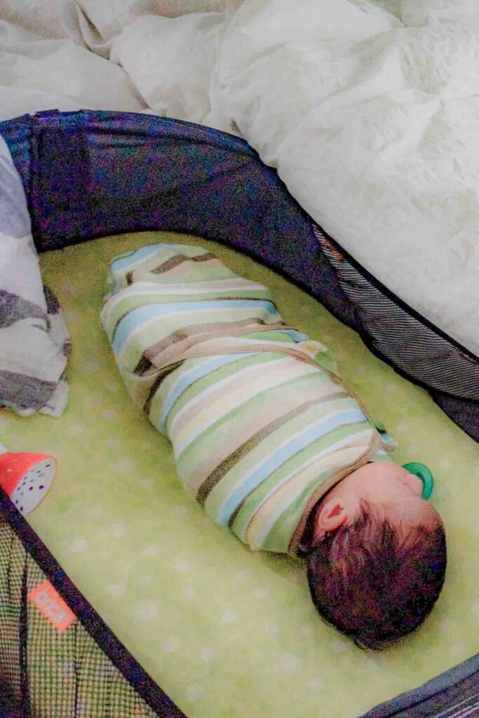 Newborn swaddled burrito style in a bright green, blue and brown SwaddleMe sleeping in a Brica travel bassinet highlighting safe baby sleep on the go and portable sleep solutions for baby during trips.