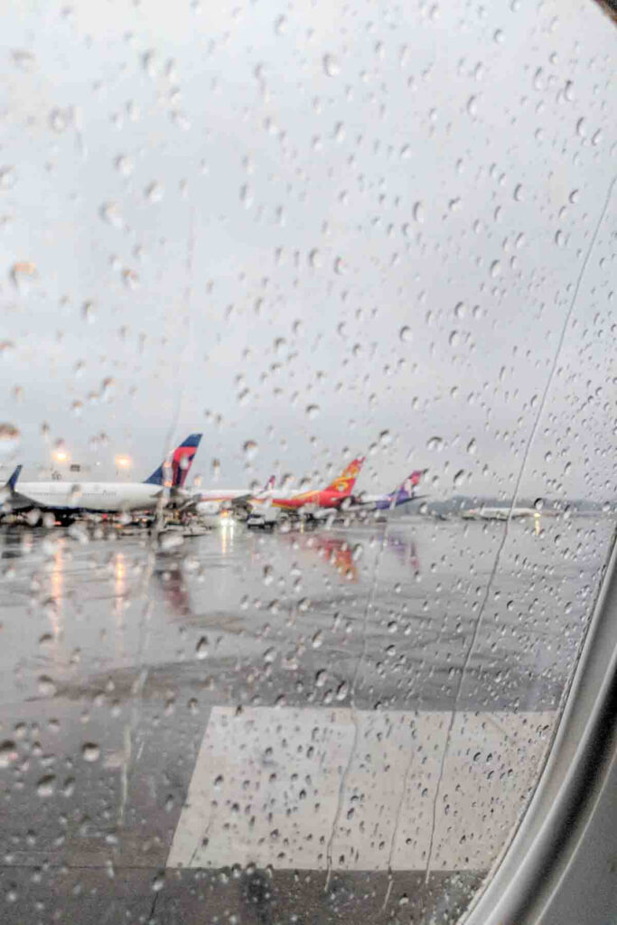 Raindrops cover an airplane window while several planes sit on a wet runway outside. 