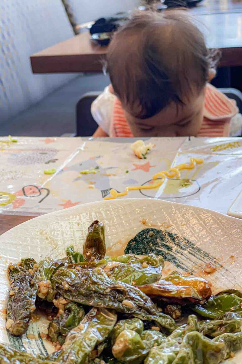 Plate of blistered shishito peppers in the foreground with a baby in a high chair looking down at her lap at dropped food behind it, highlighting the messy reality of feeding baby at a restaurant while traveling.