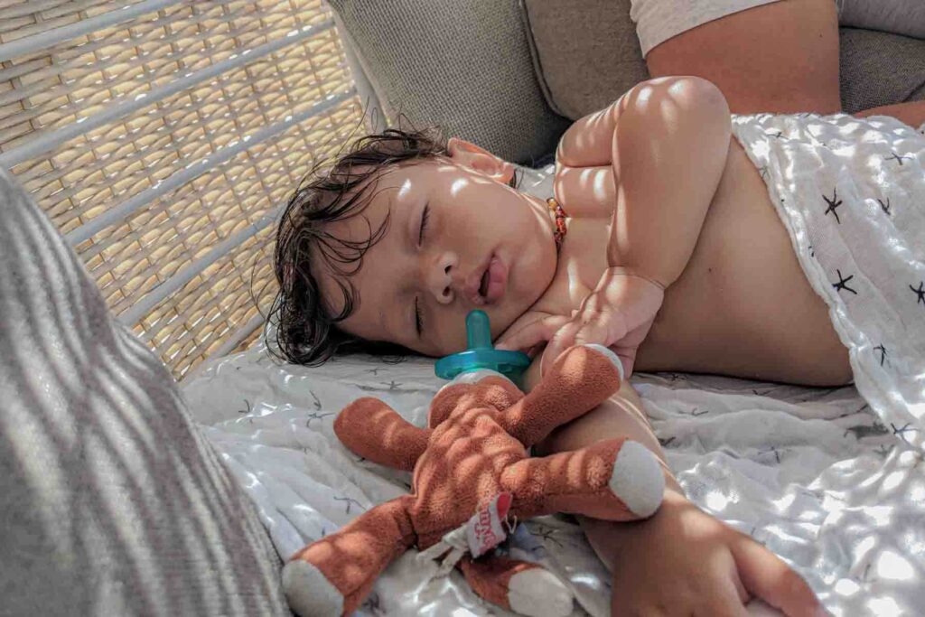 Baby sleeping with a light white muslin blanket on a cushioned daybed with a Wubbanub pacifier while sunlight filters through the woven wicker. This calm setup shows baby naps on the beach can work well.