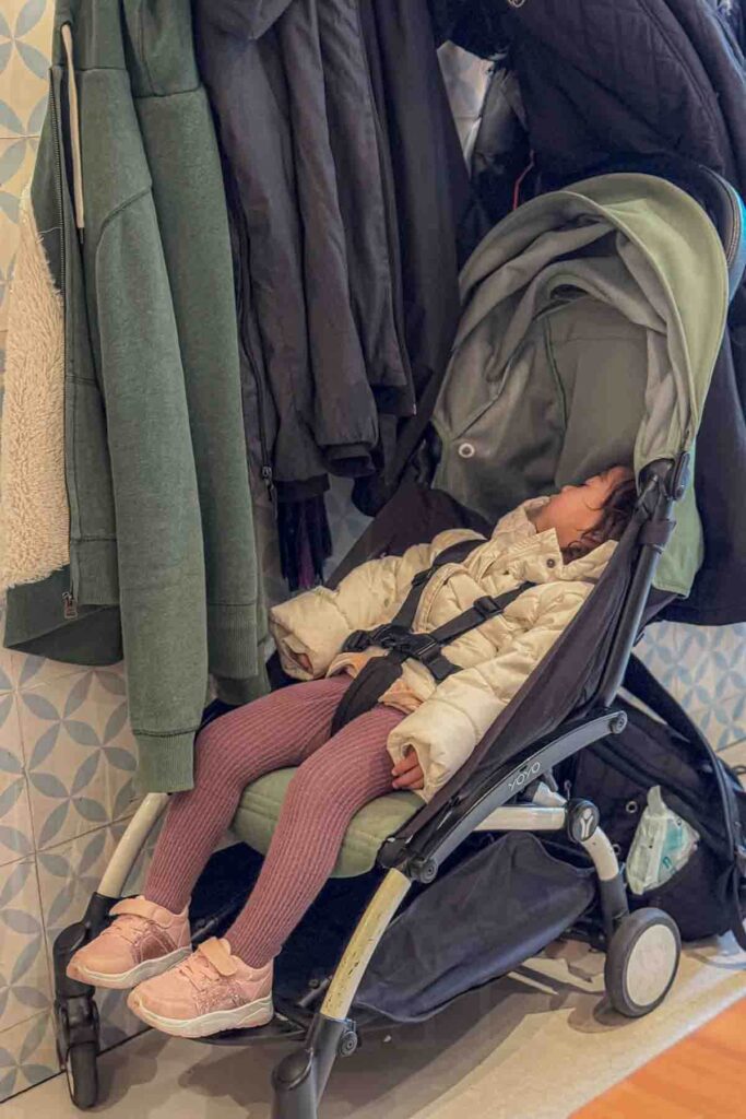 Toddler wearing a white puffy coat and pink sneakers asleep in a compact green travel stroller parked indoors beside hanging coats. This image shows how sleeping on the go while traveling works using a lightweight stroller.