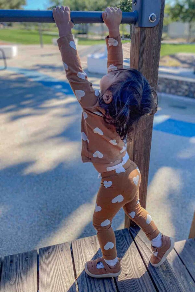 A toddler in a brown outfit with white hearts hangs from a playground bar in bright morning light. Active outdoor play  supports kids in adjusting to a new time zone by getting sunlight and movement.