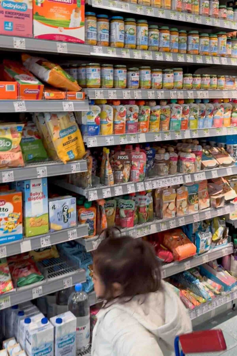 Grocery store aisle in Brasov, Romania stocked with jars and pouches of baby food and snacks while a child looks on, illustrating options for feeding a toddler in international destinations during travel.