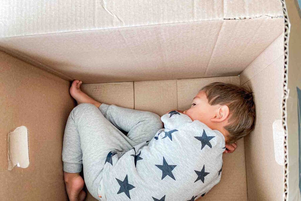 A young child curls up inside a large cardboard box wearing gray star patterned pajamas. The cozy enclosed position illustrates an overtired kid.