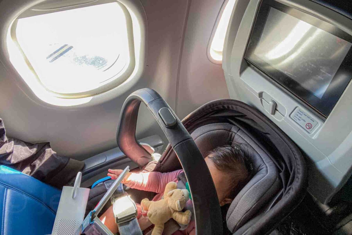 Infant resting in an Uppababy car seat by an airplane window with sunlight streaming in, illustrating flying with a baby for the first time.