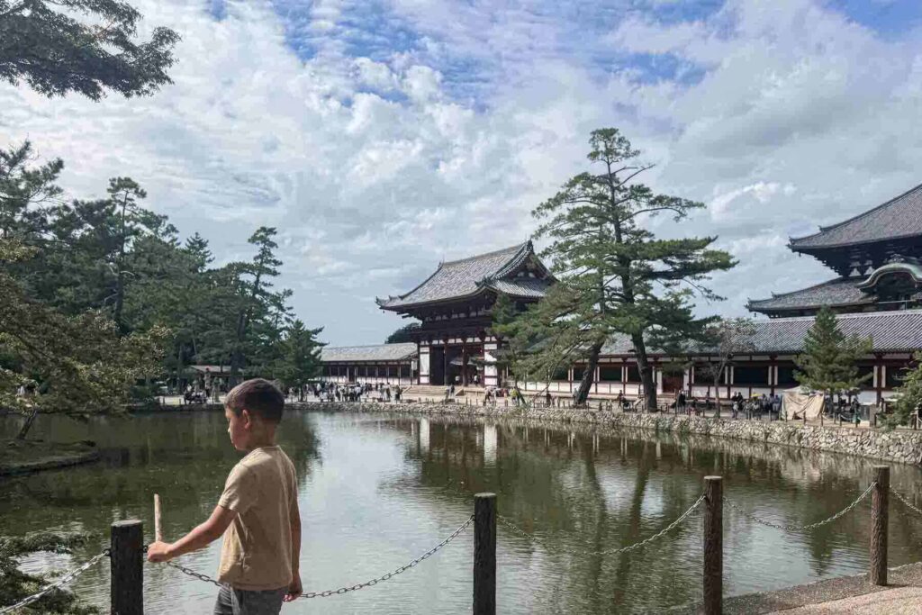 A young boy stands by a pond at Todai-ji Temple in Nara, gazing across the water toward the grand wooden gate and traditional temple buildings. This peaceful scene captures the cultural charm of one of the best day trips from Osaka with kids.