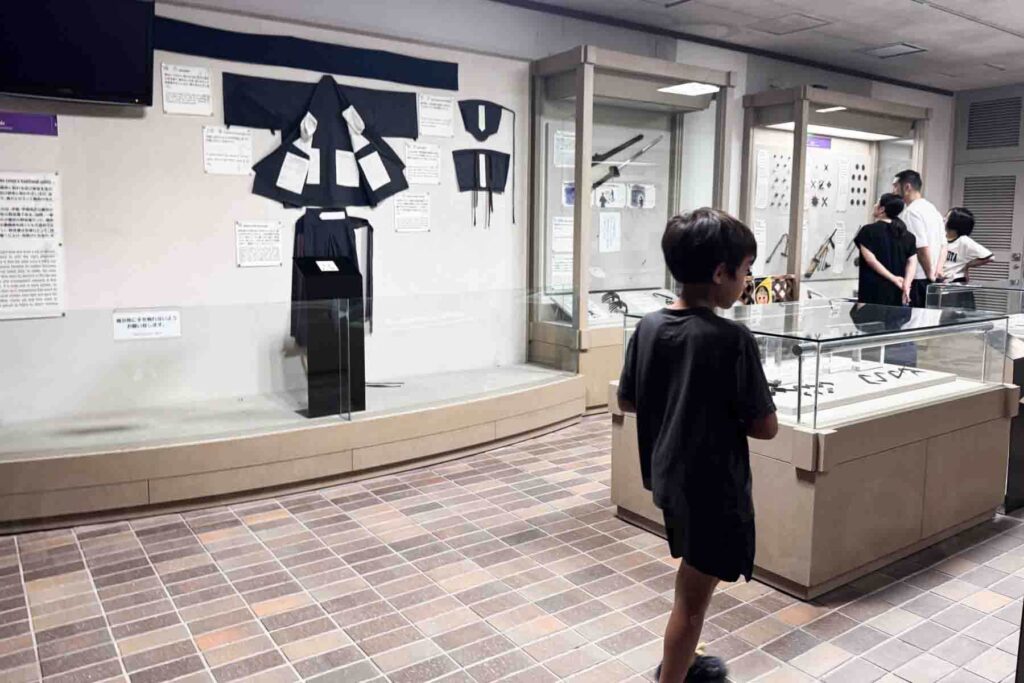 A child observes ninja-themed artifacts and clothing inside a museum, with glass display cases and traditional black and white garments on view at the Iga-ryu Ninja Museum. 