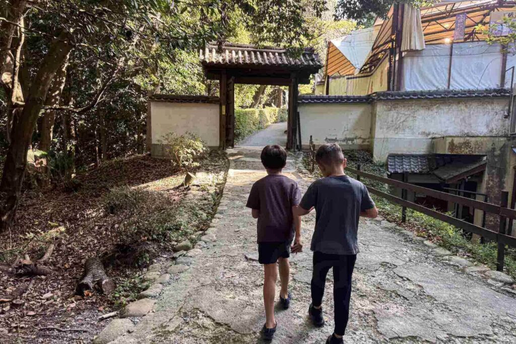 Two children walk side by side along a stone path toward a traditional Japanese gate surrounded by lush greenery on the grounds of the Iga-ryu Ninja Museum. 