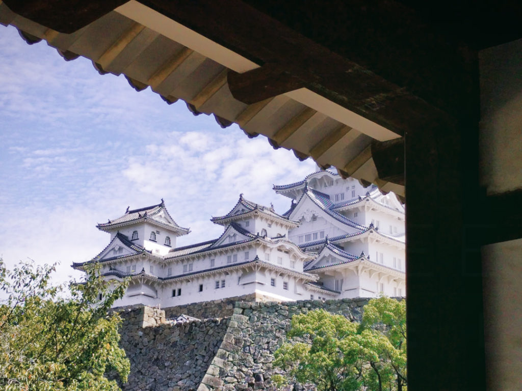 A view under a traditional awning of part of the grand white Himeji castle in Japan. The castle proudly rises against the bright blue sky above a high thick stone wall with green trees covering part of the wall.