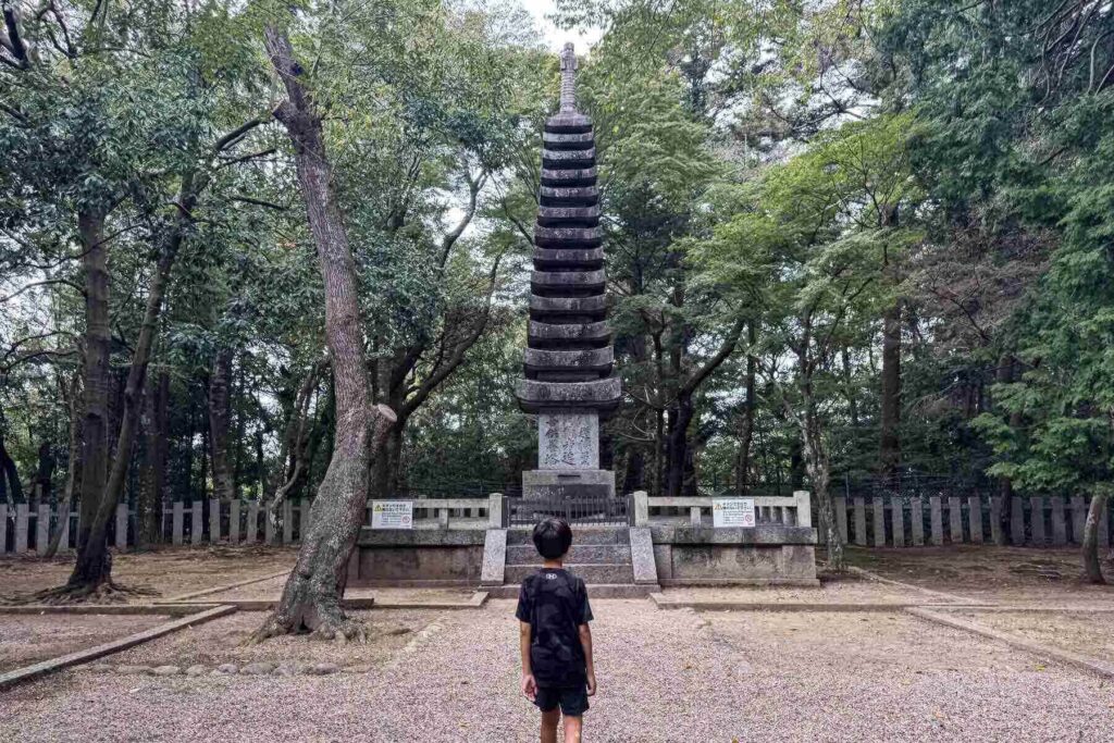 A child stands in front of a tall stone pagoda monument surrounded by forest in a peaceful area of Nara Park. 