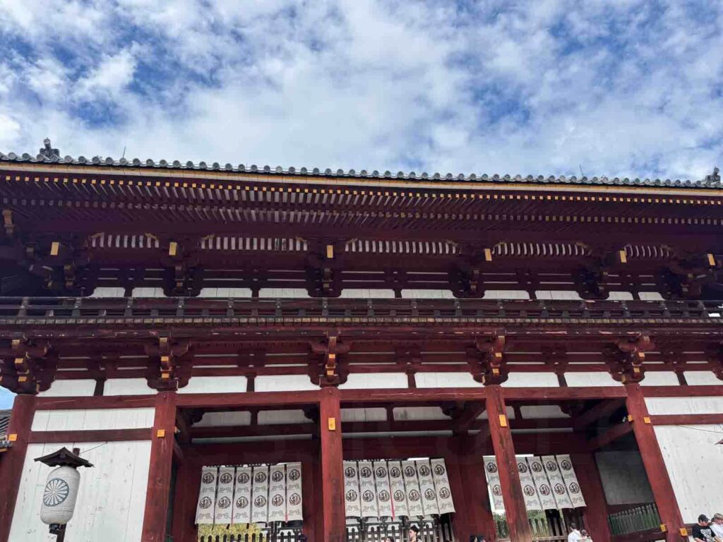 Close-up view of the vibrant red and white wooden gate of Todai-ji, a historic Japanese temple in Nara, with intricate architecture under a partly cloudy sky. 