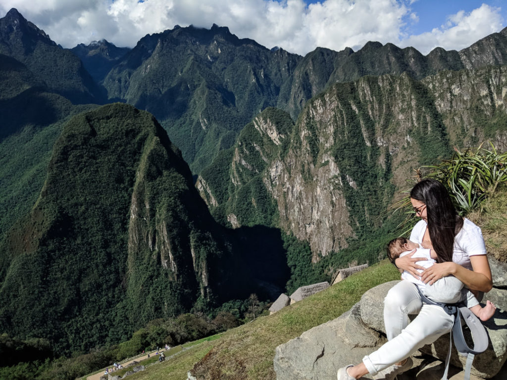 Mother perched on a rock cradling her six month old baby while he nurses peacefully with dramatic green steep mountains in the background in Peru.