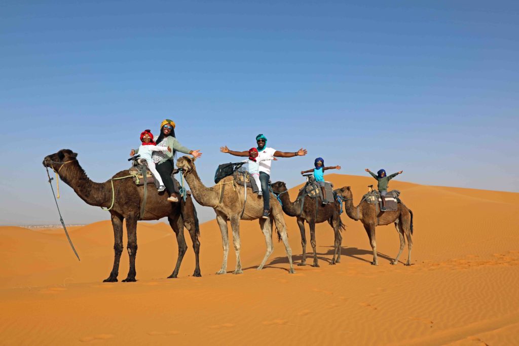 Black family traveling with kids on a camel ride through the Sahara Desert - diversity in family travel