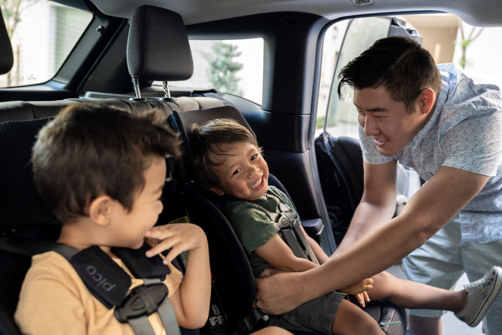 Kids smiling, dad buckling in toddler in car seat