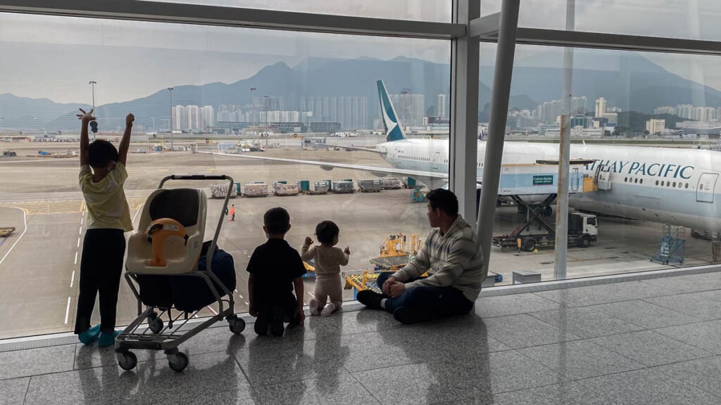 A family, including a man and three children, sits and stands by a large floor to ceiling window overlooking HKG airport tarmac with a Cathay Pacific airplane parked nearby. The children, one standing with arms raised and two sitting, seem engaged by the view, while the man sits on the floor looking at the airplane. In the background, a mountain range and the Hong Kong city skyline are visible.