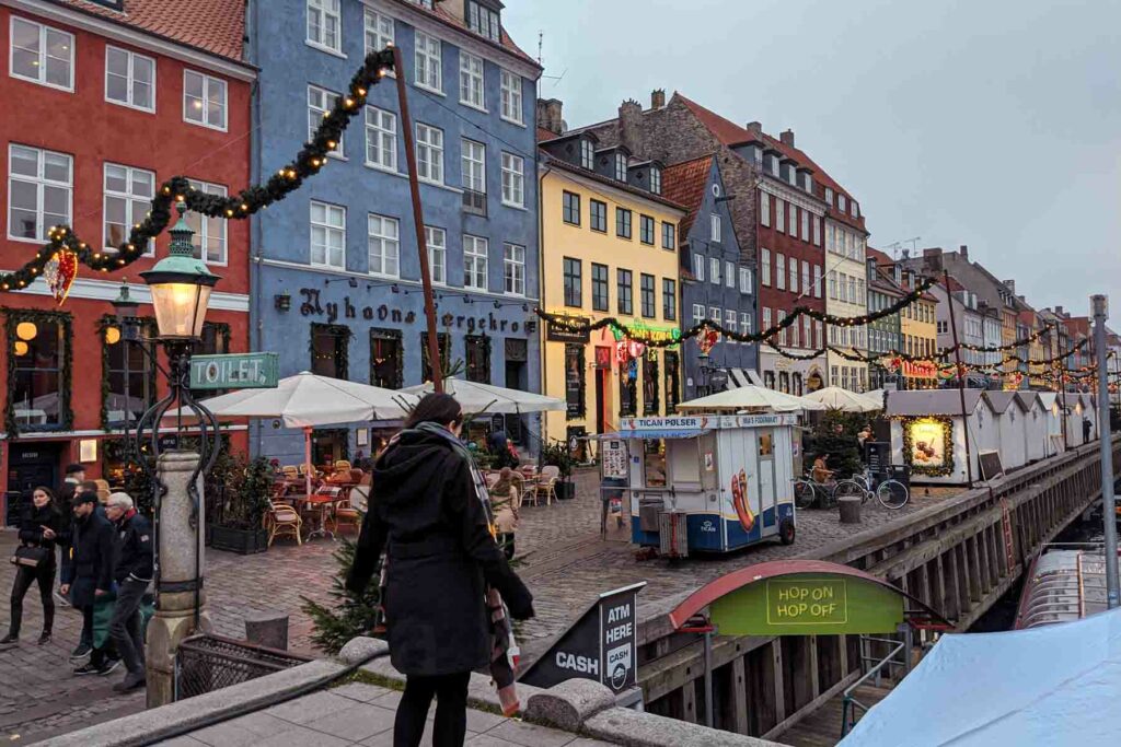 Woman wearing all black standing among colorful buildings of Nyhavn in Copenhagen, Denmark, the harbor, decorated with Christmas lights and festive decorations in winter.