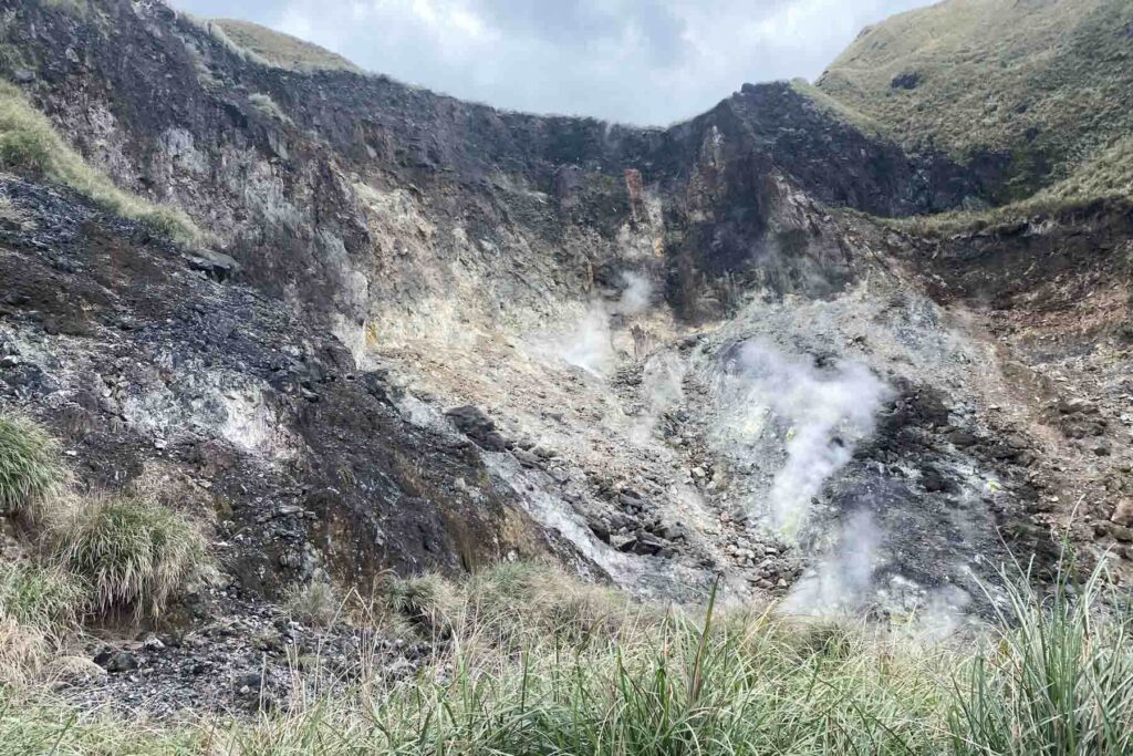 A rocky crater with exposed dark and pale earth releases plumes of steam from several vents, showing active geothermal activity at Xiaoyoukeng in Yangmingshan National Park, Taiwan. Wisps of steam rise against a backdrop of overcast skies and grassy hills.