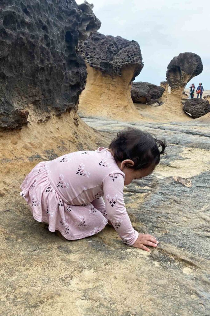 A baby in a light pink dress crouches and touches the ground among otherworldly mushroom-shaped rock formations at Yehliu Geopark in Taiwan. The unique honeycomb texture of the sandstone rocks towers behind her.