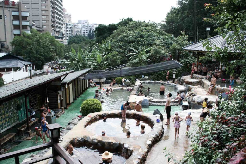 A view of public hot spring pools in Beitou, Taiwan, shows people soaking in the water near low buildings and lush greenery, with tall apartment buildings and the cityscape rising in the background.