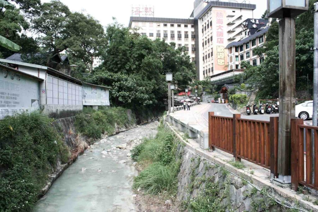 A pale, mineral-rich stream flows beside a walkway and buildings in Beitou District, Taiwan. The water is bordered by greenery and concrete retaining walls, with a backdrop of hotels and traditional-style architecture.
