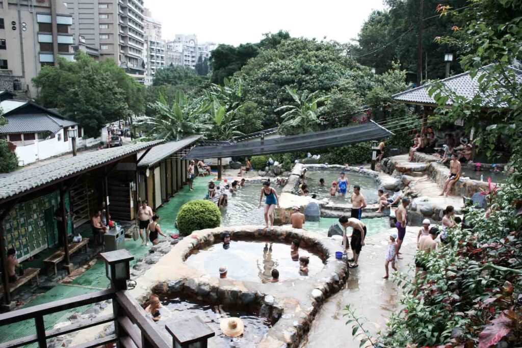 A busy scene at Beitou’s public hot springs in Taiwan shows multiple stone pools filled with bathers, surrounded by tropical plants and city buildings. Swimmers of all ages relax in the steaming waters under a canopy and alongside shaded seating areas.