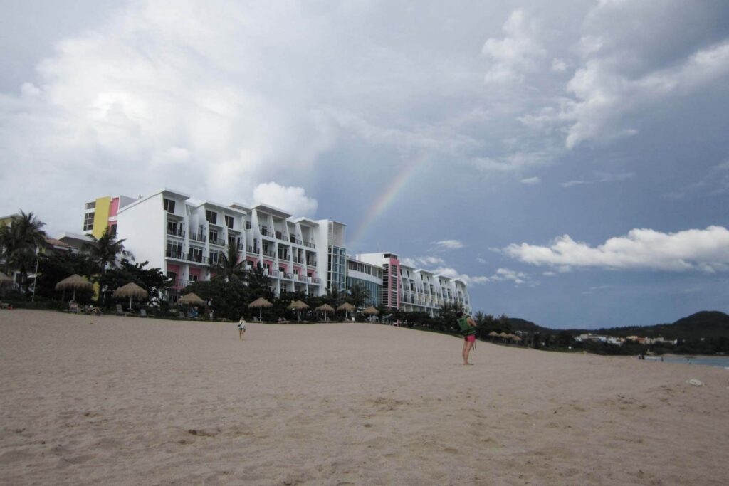 A wide sandy beach stretches out in front of the white and pink buildings of Chateau Beach Resort in Kenting, Taiwan, with a faint rainbow arcing through the cloudy sky. Palm trees and beach umbrellas line the edge of the hotel.