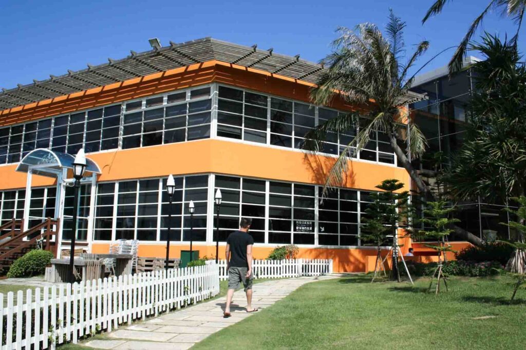 A person walks along a stone path bordered by a white picket fence toward an orange and glass building at Chateau Beach Resort in Kenting, Taiwan. Palm trees sway in the breeze under a bright blue sky.