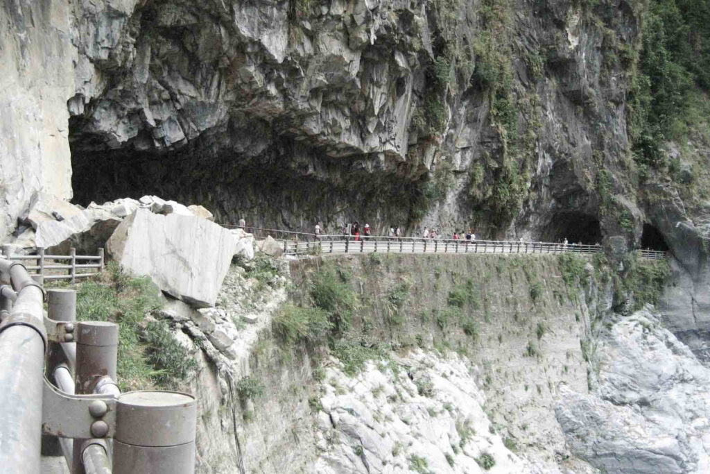 A narrow walkway hugs the steep cliffside in Taroko National Park, Taiwan, passing through rocky tunnels carved into the marble walls. Visitors walk along the protected path above a deep gorge.