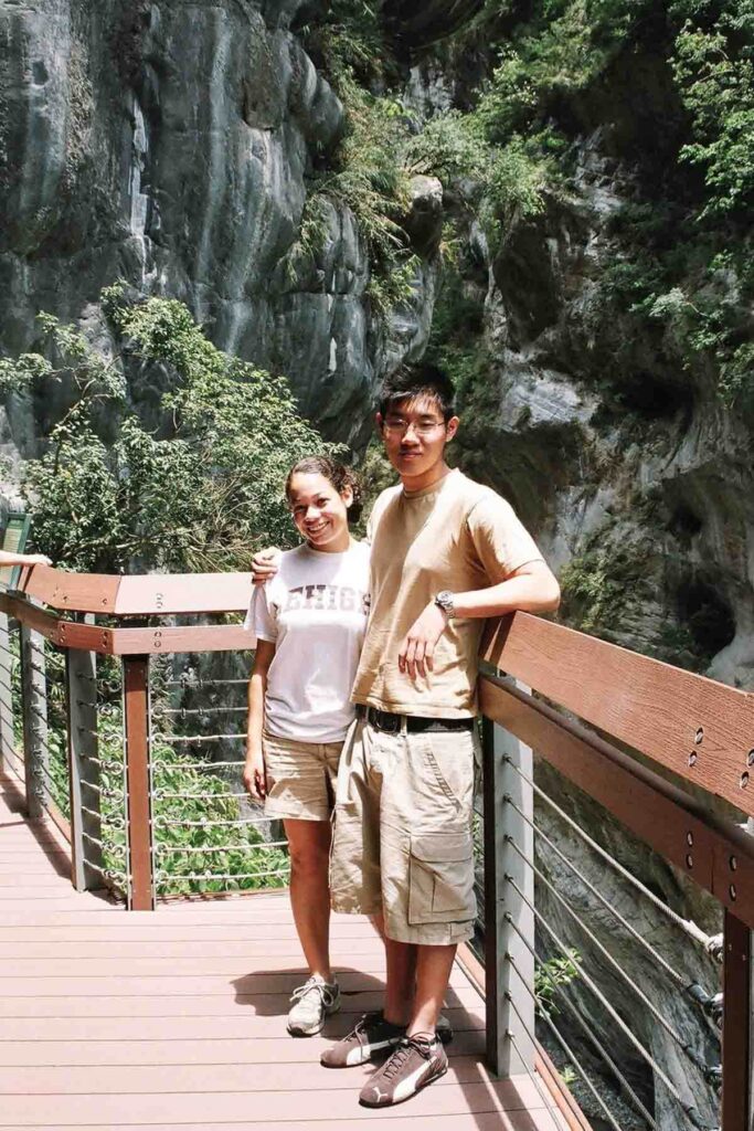 A smiling young couple poses on a wooden deck railing at Taroko Gorge in Taiwan, surrounded by towering marble cliffs and lush greenery. The man is leaning is elbow on the railing while the woman stands close beside him.
