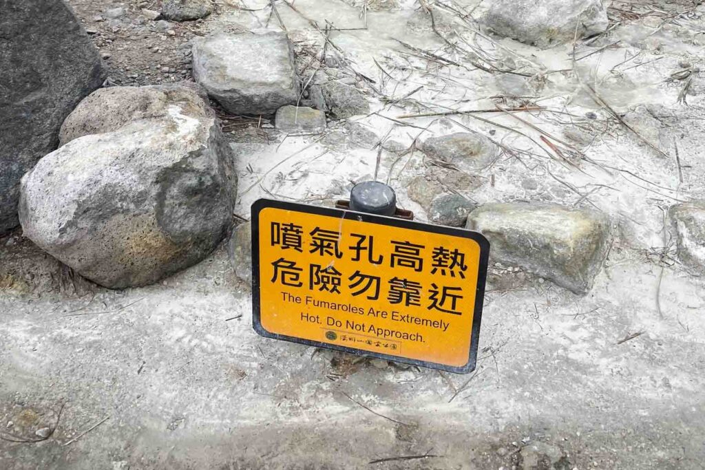 A yellow caution sign surrounded by rocks warns, “The Fumaroles Are Extremely Hot. Do Not Approach,” in English and Chinese. The ground is white and cracked, indicating geothermal activity in Xiaoyoukeng Recreation Area at Yangmingshan National Park.
