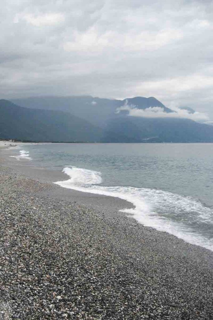 A moody shoreline view of Qixingtan Beach in Hualien, Taiwan, shows gentle waves lapping against a wide pebble beach with misty mountains looming under a cloudy sky in the background.