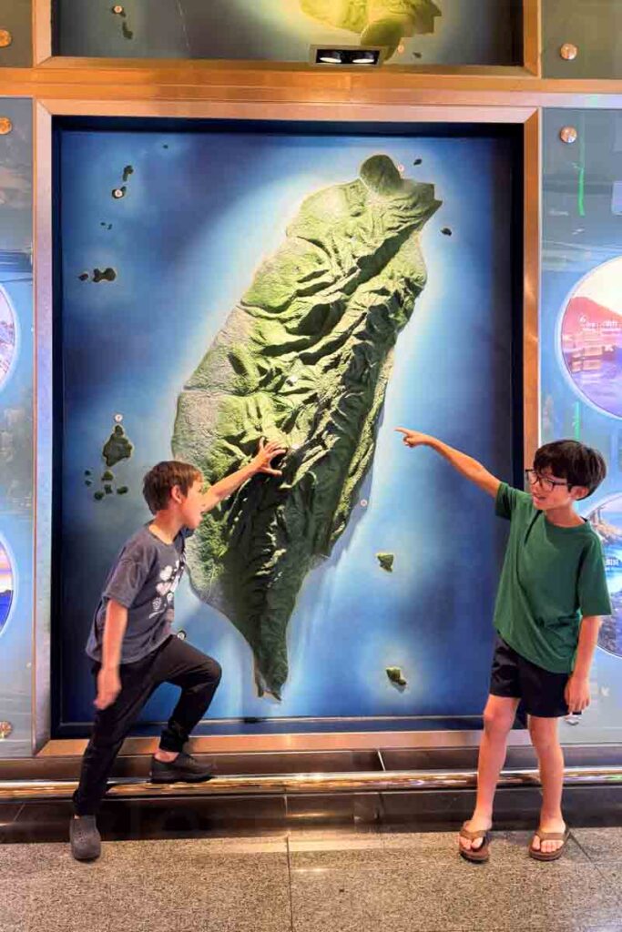 Two boys stand in front of a large topographical map of Taiwan in the Taoyuan International Airport, one pointing enthusiastically at the island’s rugged mountain spine while the other gestures across from the opposite side. The map’s green elevation contrasts against the surrounding blue ocean display.