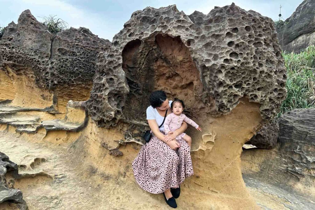 A mother in a white top and long spotted skirt sits on a honeycomb-textured rock at Yehliu Geopark in Taiwan, holding her daughter in a light pink outfit. The unique mushroom-shaped rock formation creates a cozy natural alcove around them.