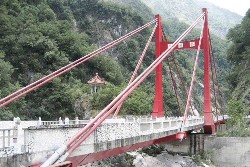 A bright red suspension bridge with bold cables spans a river in Taroko National Park, Taiwan. A traditional Chinese pavilion peeks through the dense greenery on a nearby cliffside.