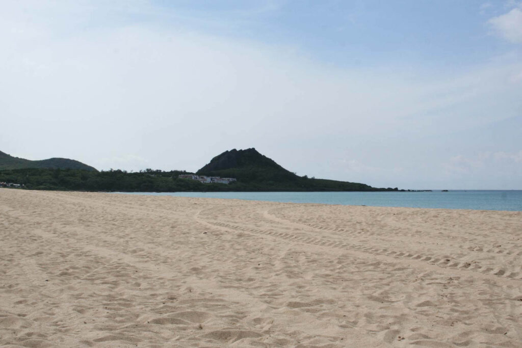 A wide, white sandy beach in Kenting, Taiwan stretches toward calm blue waters with a small deep green hill rising in the distance.