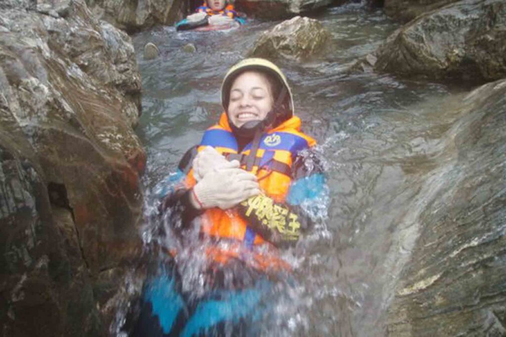 A woman in full canyoning gear—including helmet, gloves, and life vest—smiles with eyes closed as she slides down a smooth rock water chute in a narrow gorge in Hualien County, Taiwan, surrounded by rushing stream water.