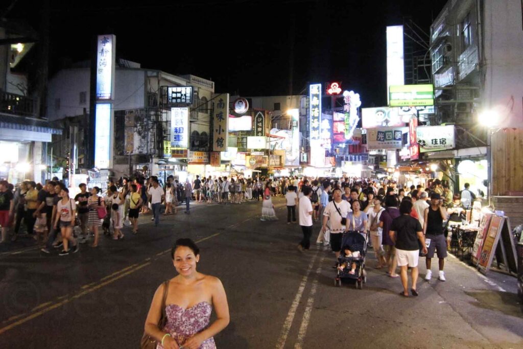 A lively night market scene in Kenting, Taiwan features neon signs, bustling crowds, and a woman in a floral dress smiling in the foreground.