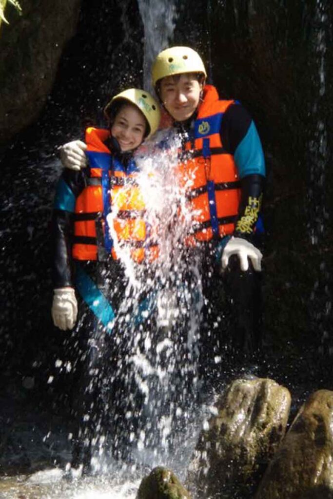 A young couple in bright orange life vests and helmets stands side-by-side under a rushing waterfall, grinning with arms around each other during a canyoning adventure in Hualien County, Taiwan.